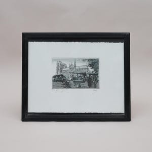 Pode incluir: Uma impressão emoldurada em preto apresentando uma gravura em preto e branco da Catedral de Notre Dame em Paris com vendedores de rua em primeiro plano. A obra está centrada num passe-partout branco com borda irregular, em moldura preta.