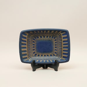 May include: A rectangular ceramic dish featuring a deep blue center and a border of radiating tan lines. The dish is supported by a black plastic display stand, showcasing a geometric pattern that extends from the solid blue base to the edges.
