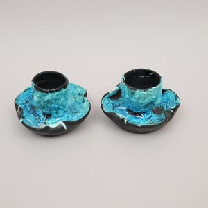 May include: Two ceramic candle holders featuring a black base with a textured, crater-like surface. The interior and upper edges are finished in a vibrant, mottled turquoise and white glaze that drips down onto the dark, uneven outer rim.