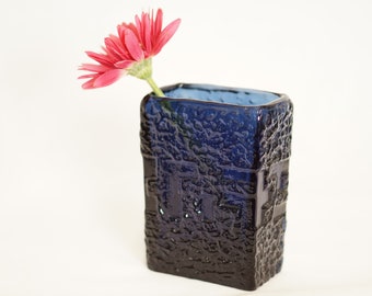 May include: A dark blue textured vase with a square shape, holding a pink gerbera daisy. The vase has a rough, patterned surface and a lighter blue interior. The flower's stem is green.