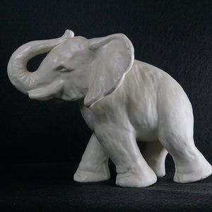 Puede incluir: Una figurita de elefante de cerámica blanca con la trompa levantada en el aire. El elefante está de pie sobre una superficie negra.