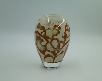 May include: A small, oval-shaped glass vase with a white background and a floral design in shades of orange and brown.