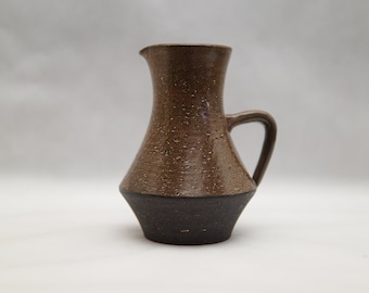 May include: A small brown ceramic pitcher with a speckled glaze and a dark brown base. The pitcher has a rounded body and a single handle.