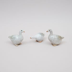 May include: Three small ceramic goose figurines with a glossy white glaze and subtle grey-toned markings. Each bird features a light orange beak and feet, with one goose positioned in the center facing downward while the other two stand upright.