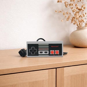 Könnte beinhalten: Ein rechteckiger, grauer und schwarzer Videospiel-Controller mit einem Steuerkreuz, zwei roten runden Tasten mit der Beschriftung A und B, zwei grauen ovalen Tasten mit der Beschriftung SELECT und START sowie dem roten Nintendo-Logo und Kabel.