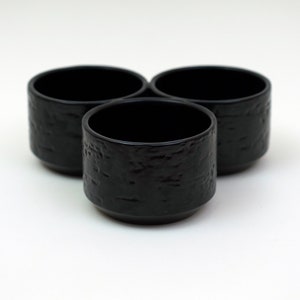 Puede incluir: Tres tazas de cerámica negra con una superficie texturizada. Las tazas están dispuestas en forma triangular.