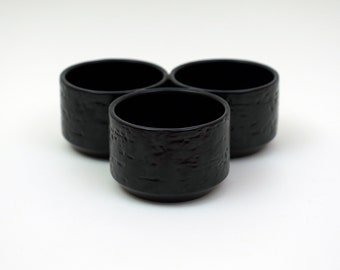May include: Three black ceramic cups with a textured surface. The cups are arranged in a triangular shape.
