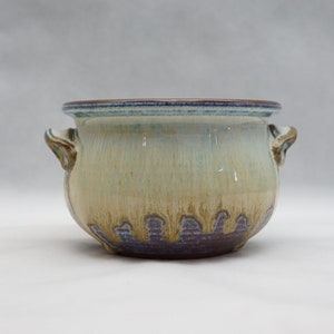 Op de afbeelding: Een keramische plantenpot met twee handvatten, met een lichtbruine, blauwe en witte glazuur. De pot heeft een ronde vorm en een licht uitlopende rand.