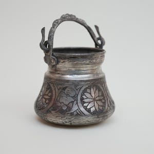 May include: A small, silver-coloured metal bucket with an arched handle. The bucket features intricate floral and leaf patterns etched into its surface. The handle is attached with decorative brackets.