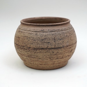 May include: A round, brown ceramic pot with a textured surface. The pot has a wide opening and a slightly rounded bottom.