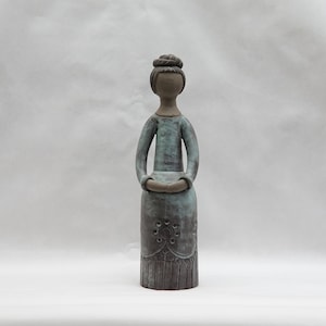 May include: A ceramic figurine of a woman with a blue and brown glaze. She is standing with her hands clasped in front of her.