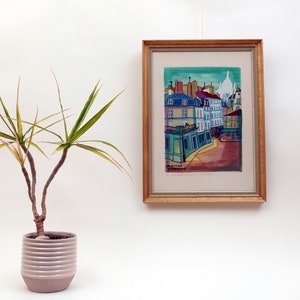 May include: A framed watercolor painting of a cityscape with colorful buildings and a blue sky. The painting is in a wooden frame with a gold finish. The painting is hanging on a white wall.