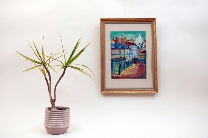 May include: A framed watercolor painting of a cityscape with colorful buildings and a blue sky. The painting is in a wooden frame with a gold finish. The painting is hanging on a white wall.