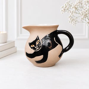 Könnte beinhalten: Ein beigefarbener Keramikkrug mit einer glänzenden schwarzen Katzenillustration, die sich um den Körper windet. Die Katze hat gelbe Augen und weiße Schnurrhaare. Der Krug hat einen geschwungenen Henkel, einen breiten Ausguss und einen runden Boden.