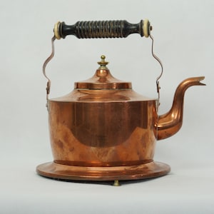May include: A copper kettle with a wooden handle and a brass knob on the lid. The kettle sits on a copper base with three legs.