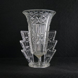 May include: A clear glass vase with a geometric design and a unique, tiered base. The vase is made of crystal and has a modern, art deco style.