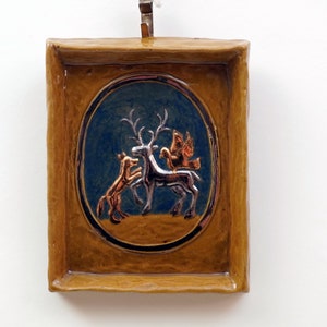 Puede incluir: Un azulejo de cerámica con un marco marrón y un óvalo azul en el centro. El óvalo presenta un ciervo plateado, un pájaro plateado y un zorro marrón.