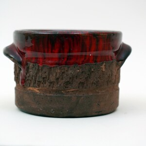 May include: A small, brown ceramic pot with a red glaze. The pot has a rough, textured surface and a slightly rounded shape.