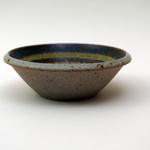 Op de afbeelding: Een kleine, grijze keramische kom met een bruine en groene rand. De kom heeft een gespikkelde textuur.