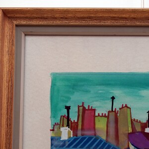 May include: A framed watercolor painting depicting a cityscape with colorful buildings and a turquoise sky. The buildings have red, green, yellow, and purple roofs. The painting is framed in a brown wood frame.