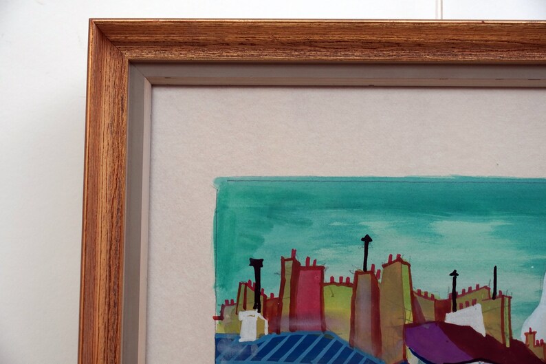 May include: A framed watercolor painting depicting a cityscape with colorful buildings and a turquoise sky. The buildings have red, green, yellow, and purple roofs. The painting is framed in a brown wood frame.