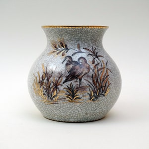 May include: A white ceramic vase with a crackled glaze and gold accents. The vase features a hand-painted design of two birds in a marsh setting.