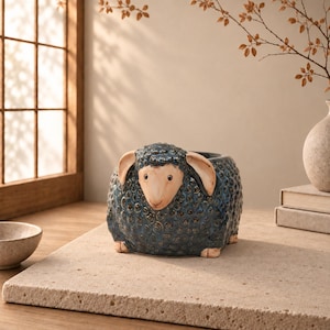 Può includere: Un vaso in ceramica a forma di pecora con un corpo in lana riccia testurizzata blu scuro e un viso liscio color beige chiaro. La pecora presenta due orecchie pendenti e piccoli piedi alla base, con un'apertura superiore per le piante.