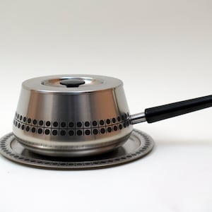 May include: A silver fondue pot with a black handle and a matching silver tray. The pot has a geometric pattern of circles and squares.