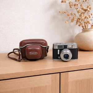 Puede incluir: Una cámara de película de 35mm Voigtländer vintage con cuerpo negro y placa superior plateada, situada junto a su funda protectora de cuero marrón a juego. La funda presenta una placa con el logotipo ovalado y el nombre Voigtländer.
