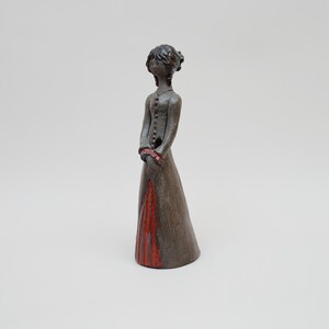 Puede incluir: Figura de cerámica de una mujer con un vestido largo. La figura es principalmente marrón con detalles rojos en la falda y las mangas. Las manos de la mujer están juntas frente a ella.
