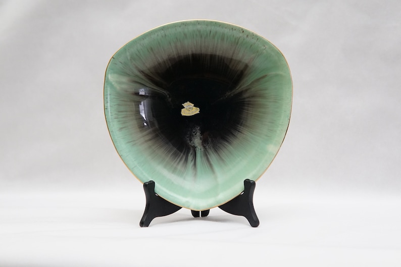 Puede incluir: Un plato de cer&aacute;mica verde y negro con un borde dorado. El plato est&aacute; sobre un soporte negro. El plato tiene un dise&ntilde;o &uacute;nico con un centro negro y verde que irradia hacia afuera.