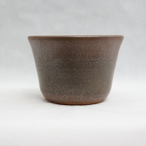 Pode incluir: Um vaso de cerâmica castanho com uma textura salpicada. O vaso tem uma borda ligeiramente alargada e uma base arredondada. A cor é um castanho suave com salpicos mais escuros, dando-lhe um aspeto rústico. Adequado para uso interno ou externo.
