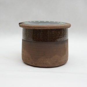 Puede incluir: Un recipiente de mantequilla de cerámica marrón con tapa. La tapa tiene un esmalte marrón oscuro con un borde blanco.