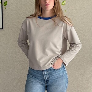 Peut inclure: Un chemisier à manches longues gris clair avec une bordure bleue autour de l'encolure. Le chemisier est fait d'un tissu doux et confortable et a une coupe décontractée.