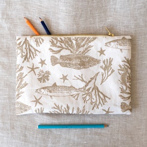 Può includere: Una pochette bianca con cerniera dorata. La pochette ha uno sfondo beige con un motivo ripetuto di pesci marroni, stelle marine e alghe.