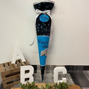 Könnte beinhalten: Ein blauer und schwarzer Schultüte mit einem weißen Band und einem Anhänger mit der Aufschrift "Schulkind 2023". Die Tüte hat einen blauen Abschnitt mit einem Cartoon-Delfin und dem Namen "Emir Yuv" in weiß.