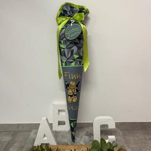 Könnte beinhalten: Ein grauer und grüner Schultüte mit einem grünen Band und einem Etikett mit der Aufschrift "Schulkid 2023". Die Tüte hat ein Bild einer Lego-Ninja-Figur und den Namen "Finn" in goldenen Lettern.