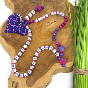 Könnte beinhalten: Eine violette und rosafarbene Alphabet-Perlenkette mit einem violetten herzförmigen Anhänger. Die Kette hat Holzperlen mit weißen Buchstaben und violetten und rosafarbenen Abstandshaltern. Die Kette liegt auf einer Holzoberfläche.