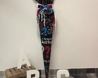 Può includere: Un cono scolastico nero e blu con un nastro bianco e un'etichetta rossa che dice "Schulkind 2023". Il cono è decorato con punti interrogativi blu e rossi e lettere. Il cono è etichettato "1. Detektiv Justus".