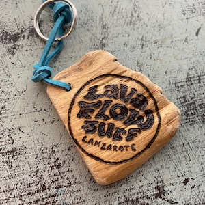 Pu&ograve; includere: Un portachiavi in legno di colore marrone chiaro e forma irregolare, caratterizzato da un disegno circolare pirografato con il testo "LAVA FLOW SURF LANZAROTE". Un cordino in camoscio blu collega il legno a un anello metallico argentato.