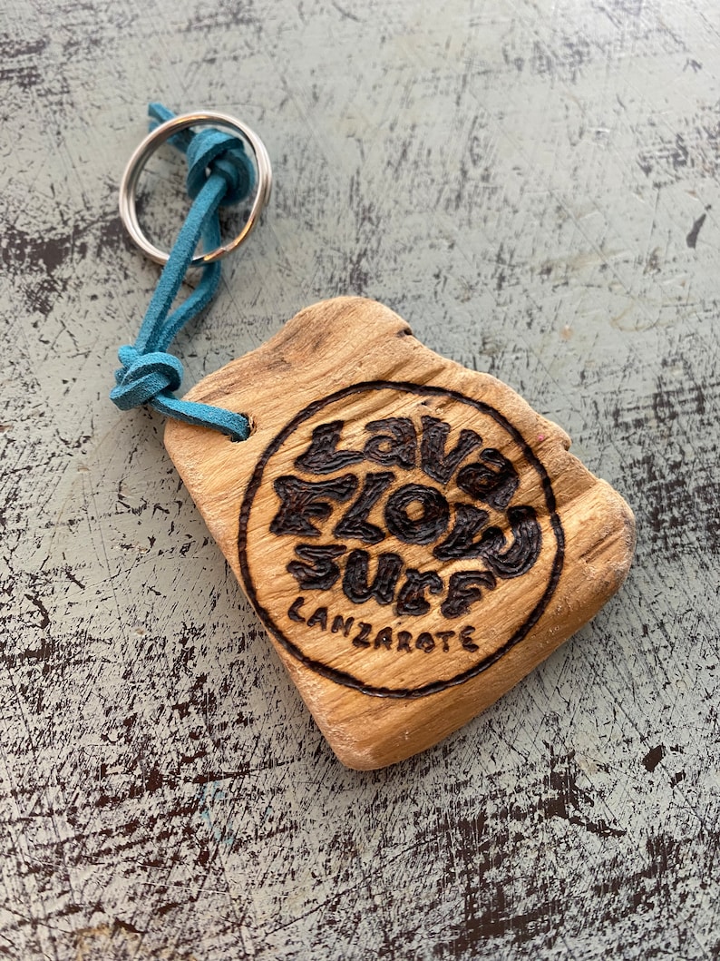 Pu&ograve; includere: Un portachiavi in legno di colore marrone chiaro e forma irregolare, caratterizzato da un disegno circolare pirografato con il testo "LAVA FLOW SURF LANZAROTE". Un cordino in camoscio blu collega il legno a un anello metallico argentato.