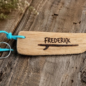 Pu&ograve; includere: Un portachiavi in legno di deriva chiaro di forma rettangolare con il nome Frederick inciso a fuoco sopra un grafico di una tavola da surf. Un cordino in pelle turchese brillante collega il legno a un anello portachiavi circolare in metallo.
