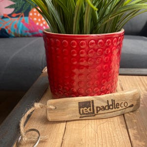 Pu&ograve; includere: Un portachiavi rettangolare in legno galleggiante di colore chiaro con il testo "red paddleco" impresso sulla superficie. Un pezzo di spago di juta naturale &egrave; infilato in un foro sul lato sinistro, collegato a un anello portachiavi circolare in metallo argentato.