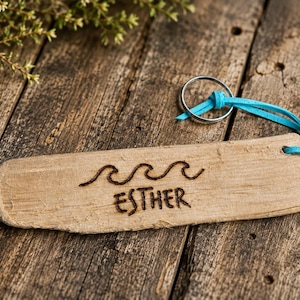 Pu&ograve; includere: Un portachiavi rettangolare in legno di deriva color marrone chiaro con un disegno pirografato di tre onde oceaniche e il nome ESTHER. Un cordino in pelle turchese brillante collega il legno a un anello portachiavi circolare in metallo.