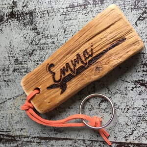 Pu&ograve; includere: Un pezzo rettangolare di legno galleggiante marrone chiaro con il nome Emma inciso a fuoco sulla superficie. Un cordino in camoscio arancione &egrave; infilato in un foro nel legno e fissato a un portachiavi circolare in metallo argentato.