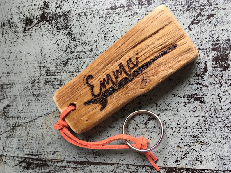 Pu&ograve; includere: Un pezzo rettangolare di legno galleggiante marrone chiaro con il nome Emma inciso a fuoco sulla superficie. Un cordino in camoscio arancione &egrave; infilato in un foro nel legno e fissato a un portachiavi circolare in metallo argentato.