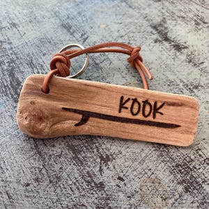 Pu&ograve; includere: Un portachiavi rettangolare in legno galleggiante color marrone chiaro con un disegno pirografato di una tavola da surf e la parola KOOK. Un cordino in pelle beige &egrave; infilato in un foro nel legno e fissato a un anello portachiavi in metallo.
