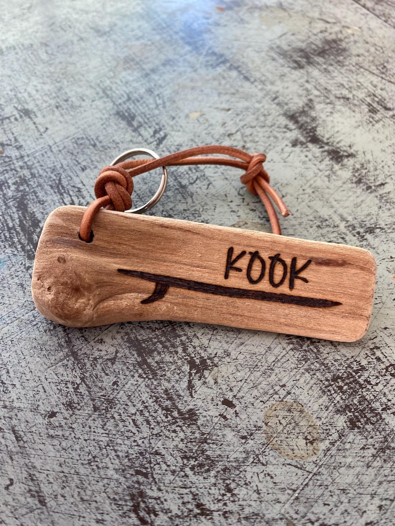 Pu&ograve; includere: Un portachiavi rettangolare in legno galleggiante color marrone chiaro con un disegno pirografato di una tavola da surf e la parola KOOK. Un cordino in pelle beige &egrave; infilato in un foro nel legno e fissato a un anello portachiavi in metallo.