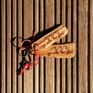 Pu&ograve; includere: Due portachiavi rettangolari in legno con un motivo a onde inciso e il testo "go with the flow". Ogni portachiavi &egrave; fissato a un anello in metallo argentato tramite un sottile cordino arancione o rosso brillante, con venature del legno.