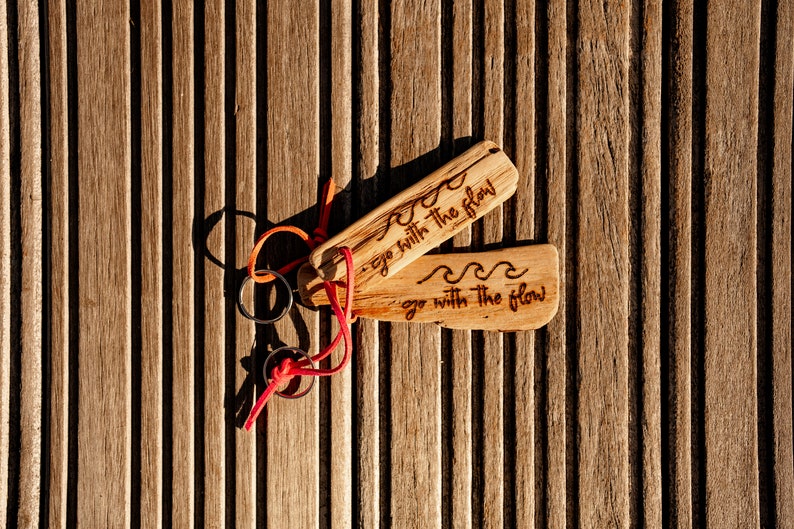 Pu&ograve; includere: Due portachiavi rettangolari in legno con un motivo a onde inciso e il testo "go with the flow". Ogni portachiavi &egrave; fissato a un anello in metallo argentato tramite un sottile cordino arancione o rosso brillante, con venature del legno.